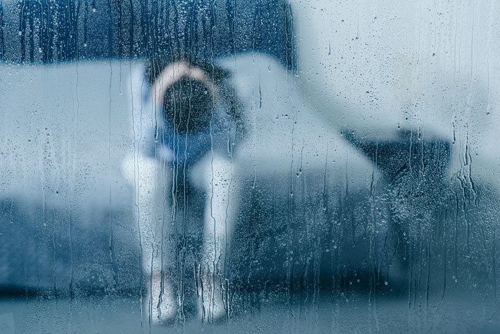 depressed woman sitting on bed, seen through rainy window - co-occurring disorders
