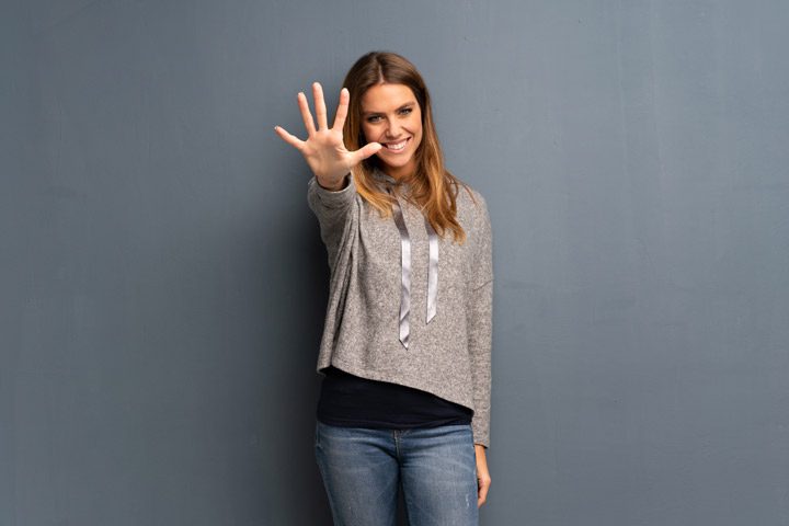 smiling woman holding out hand to gesture the number five on grey background - residential treatment